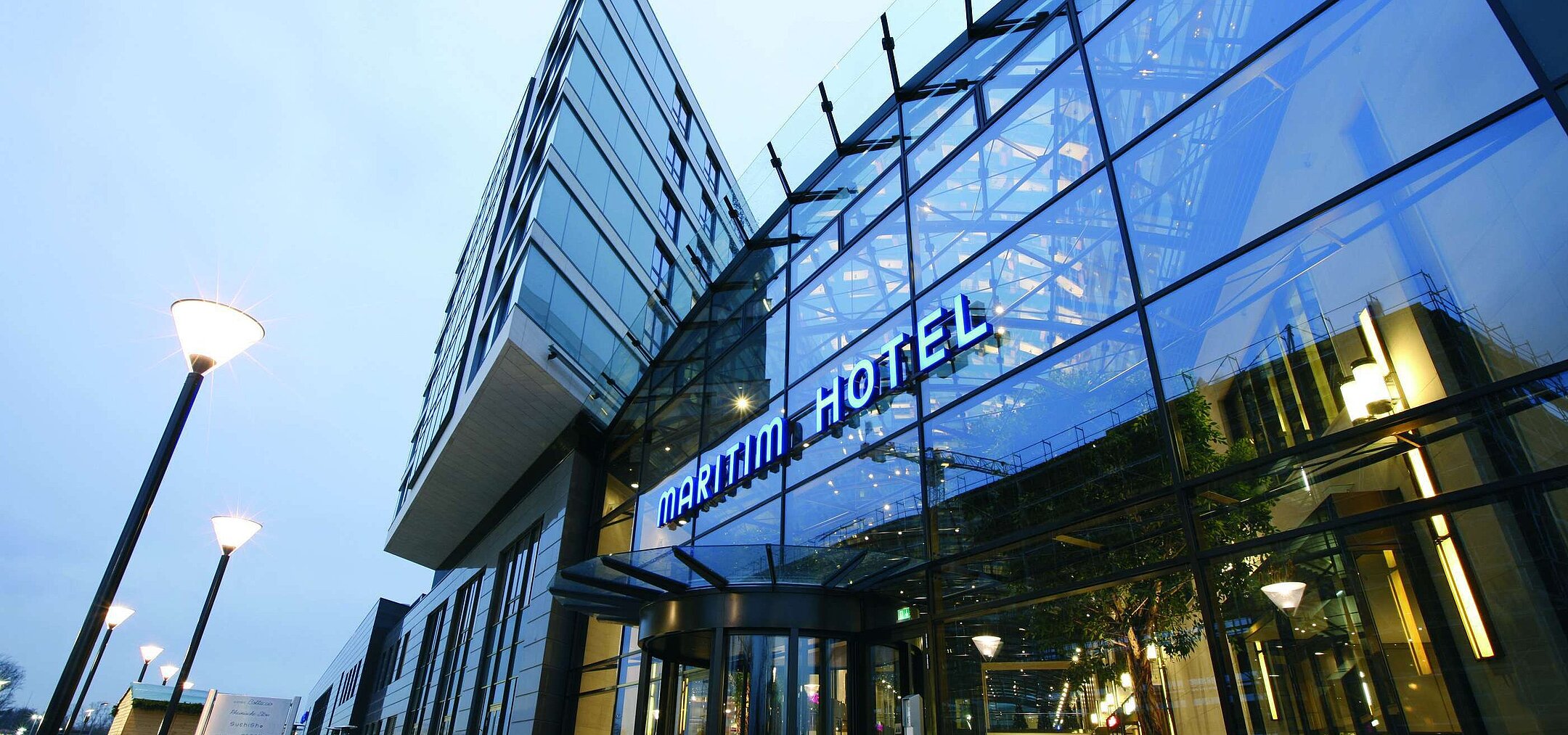 "Moderne Außenansicht des Maritim Hotel Düsseldorf mit gläserner Fassade und leuchtendem Hotellogo bei Dämmerung.