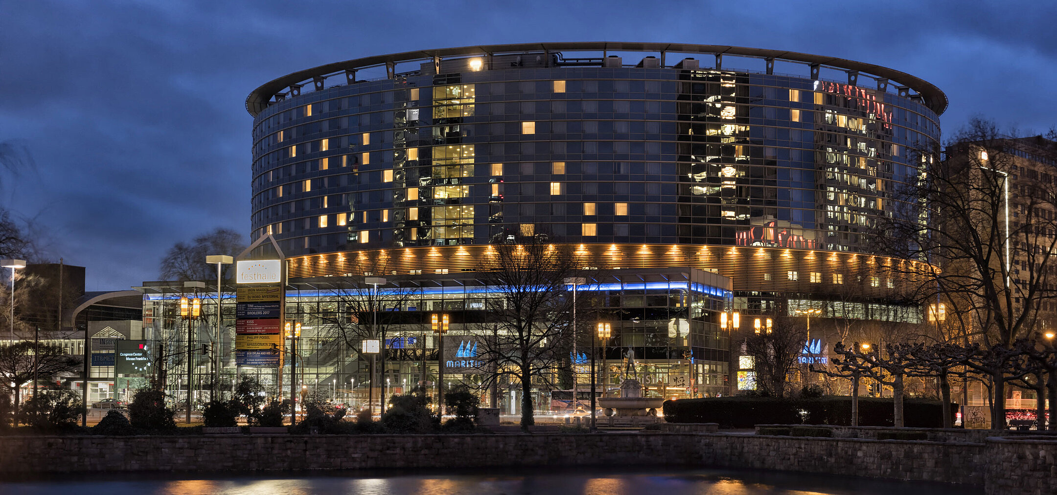 Maritim Hotel Frankfurt bei Nacht mit beleuchteter Glasfassade und Spiegelung im ruhigen Teich davor.