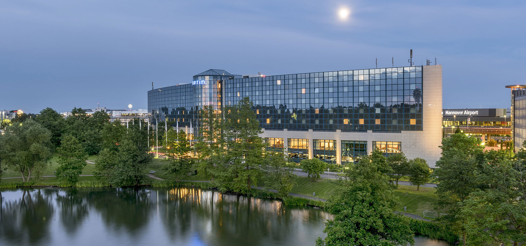 Beleuchtetes Maritim Hotel Hannover mit Glasfassade und Seespiegelung in der Abenddämmerung.