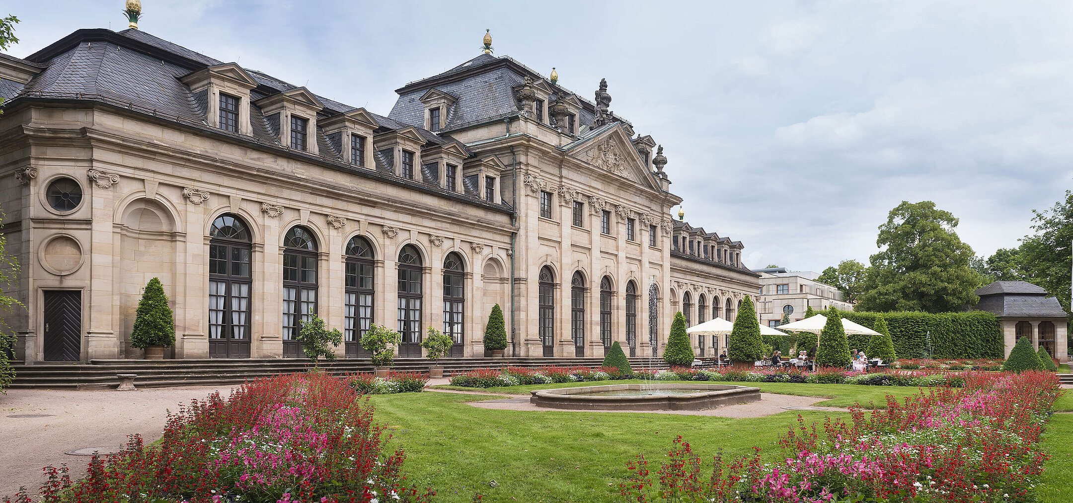 Die historische Orangerie in Fulda mit barocker Fassade, Treppe und blühendem Garten im Vordergrund.
