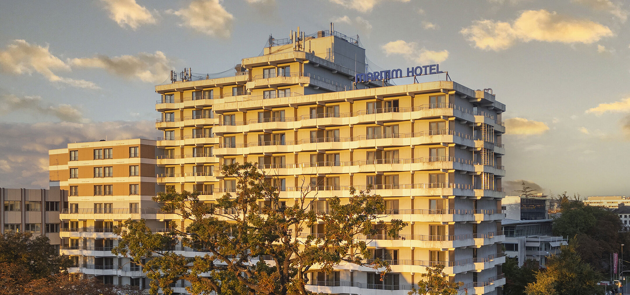 Außenansicht des Maritim Hotel Darmstadt bei Sonnenuntergang mit moderner Architektur und Bäumen im Vordergrund.