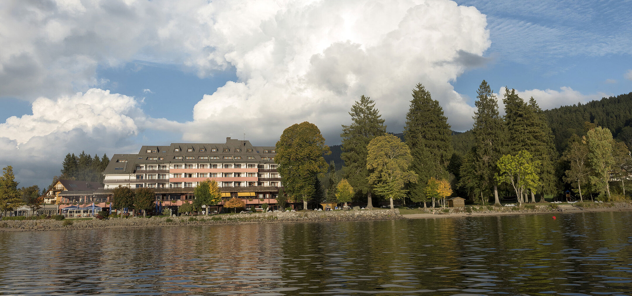 Außenansicht des Maritim Hotel Titisee mit Bäumen und Seeufer bei blauem Himmel und weißen Wolken