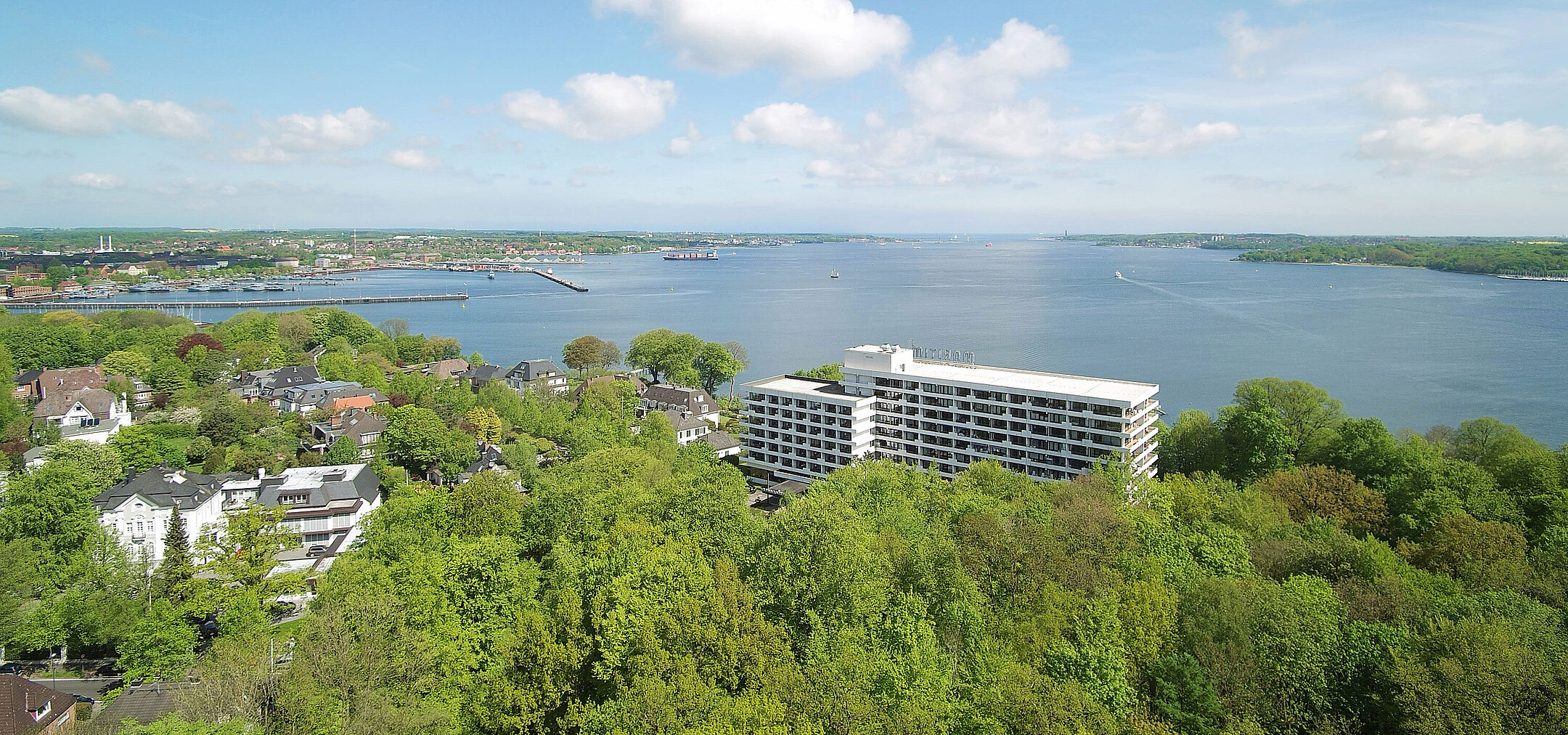 Luftaufnahme des Maritim Hotels Kiel, umgeben von grüner Landschaft und der Kieler Förde, bei strahlend blauem Himmel.